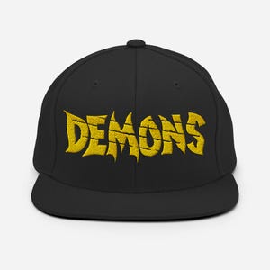 Puede incluir: Gorra de béisbol negra con la palabra "DEMONS" bordada en amarillo, con letras estilizadas. La gorra tiene una visera plana y una corona redondeada. El texto es en negrita y texturizado.