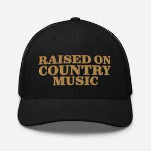 May include: Black trucker hat with the embroidered text "RAISED ON COUNTRY MUSIC" in a gold color. The hat has a mesh back and a curved brim. A classic accessory for country music fans.