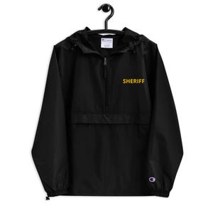 May include: Black Champion windbreaker jacket with a gold embroidered "SHERIFF" on the front. The jacket has a zipper closure and a hood.