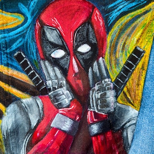 May include: A denim jacket featuring a hand-painted image of Deadpool. The artwork depicts Deadpool's red and black mask, with his hands near his face, and two swords. The background has a colorful, artistic style.