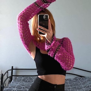 Puede incluir: Un bolero de ganchillo rosa brillante con mangas largas. Las mangas tienen ribetes negros en los puños y los hombros. La prenda se lleva sobre un top corto negro y se combina con pantalones negros. El estilo es moderno y a la moda.