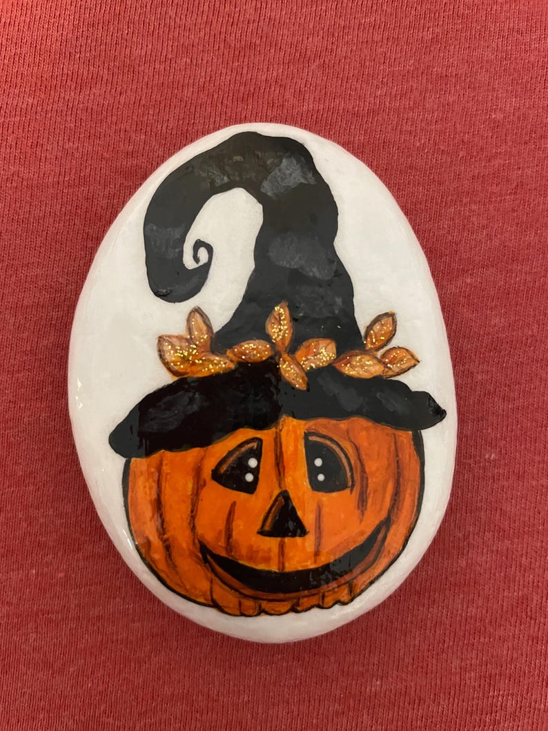 May include: A hand-painted rock with a smiling jack-o'-lantern face wearing a black witch hat with gold glitter accents.