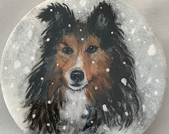 May include: A painted round ornament featuring a brown and black dog with white markings. The dog is depicted in a snowy scene with white paint splatters representing falling snow.
