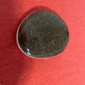 May include: A smooth, oval-shaped black stone with a white rim. The stone has a speckled texture.