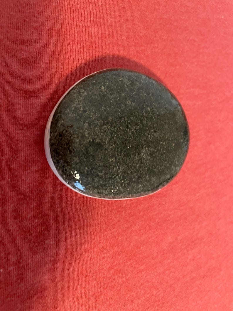 May include: A smooth, oval-shaped black stone with a white rim. The stone has a speckled texture.