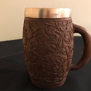 May include: A brown wooden mug with intricate floral carvings. The mug features a handle and a silver-colored rim. The detailed carvings cover the entire body of the mug, creating a textured surface. The mug is designed for beverages.