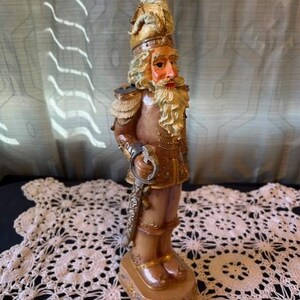 May include: A decorative nutcracker figurine in a soldier's uniform. The figurine has a detailed face with a long white beard and a gold-colored helmet. The uniform is brown and tan with gold accents. The figurine is approximately 10 inches tall.