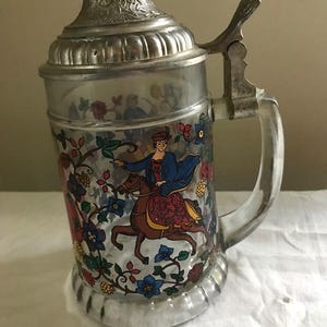 May include: A glass beer stein with a silver lid and handle. The stein features a colorful painted scene of a woman riding a horse through a floral landscape.