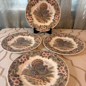 May include: A set of decorative ceramic plates featuring a turkey design. The plates have a cream-colored background with a detailed illustration of a turkey surrounded by floral and foliage borders. The plates are displayed on a table.