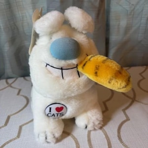 May include: A plush white cat-like toy with a blue nose and a yellow and orange beak. The toy has a black stitched smile and two white ears. A button on the toy reads "I ❤️ CATS".