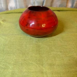 May include: A small, round ceramic bowl with a glossy finish, featuring a vibrant red color with darker, mottled black accents throughout the surface. The bowl has a wide, open top and a smooth, curved body, showcasing a deep red interior.