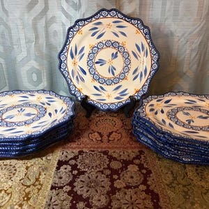 May include: A collection of ceramic plates with a blue and white floral design. The plates have a scalloped edge and a central medallion pattern. The plates are stacked in sets, with one plate displayed on a stand.