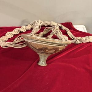 May include: A handcrafted ceramic planter with a pedestal base and a macrame plant hanger. The planter is brown with green and white accents. The macrame hanger is made of natural-colored rope. The planter is sitting on a red velvet surface.