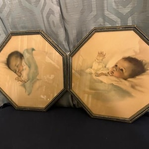 May include: Two framed vintage baby portraits. One shows a sleeping baby wrapped in a light blue blanket, the other shows a baby awake with its arm raised. Both are in octagonal frames with a gold trim.