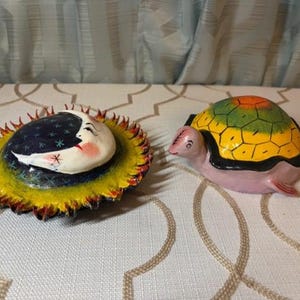 May include: Two ceramic figurines: a sun and moon face on a yellow base, and a turtle with a yellow, green, and orange shell. The sun and moon face has a white crescent moon with a sleeping face on a dark blue background with stars.
