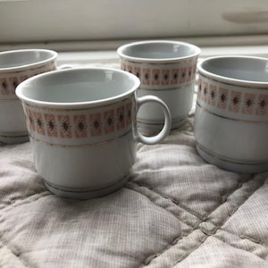 May include: Four white ceramic coffee cups with a decorative band of pink and brown geometric patterns and gold trim. Each cup has a handle and sits on a quilted surface. The cups are arranged in a group.