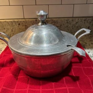 May include: A silver serving bowl with a lid and a ladle. The bowl has a round shape and a smooth surface. The lid has a knob on top. The ladle is attached to the side of the bowl.