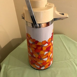 May include: Vintage cream-colored thermos with a chrome handle and spout. The lower portion is decorated with a floral pattern in shades of orange, red, and white. The thermos has a cream-colored lid and a silver band around the top.
