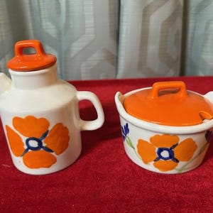 May include: A white ceramic creamer and sugar bowl set. The creamer has a handle and spout, and the sugar bowl has a lid. Both pieces are decorated with orange and blue floral designs. The lids are orange.
