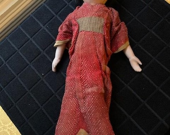 May include: A vintage Japanese doll with a painted face and black hair. The doll wears a red patterned kimono with a brown sash and gold trim. The doll is posed on a black textured surface.