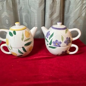 May include: Two white ceramic teapots with matching teacups. One teapot has yellow tulip designs, and the other has purple flower designs. Both have white handles and lids. The teapots are on a red surface.