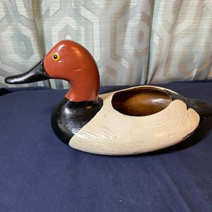 May include: A wooden duck figurine with a red head, black beak, and black and white body. The duck has a hollowed-out area on its back. The figurine is on a blue surface.