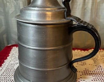 May include: A pewter beer stein with a hinged lid and a black handle. The cylindrical body features horizontal bands. The lid has a rounded top and a decorative handle. The stein sits on a white lace tablecloth.