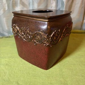 May include: A square tissue box cover featuring a deep reddish-brown finish with a textured surface. The upper section is adorned with a raised, metallic bronze-colored band displaying an intricate, swirling vine and leaf relief pattern.
