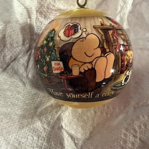 May include: A round Christmas ornament with a cartoon illustration of a character relaxing by a fireplace. The ornament has a gold cap and the text "Have yourself a cozy..." is visible.