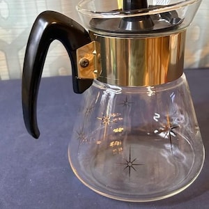 May include: A clear glass coffee carafe featuring a gold-tone metal band around the neck and a black plastic handle. The glass body is decorated with black starburst patterns. It includes a black plastic lid with a central knob component.