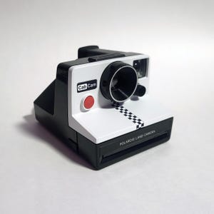 Peut inclure: Un appareil photo Polaroid Land noir et blanc avec un bouton rouge et un motif à carreaux. L'appareil photo porte l'inscription "CabCam" sur le devant.