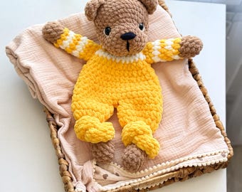 May include: A crocheted brown bear wearing a yellow crocheted jumpsuit with white accents. The bear is lying on a pink and white muslin blanket in a wicker basket.