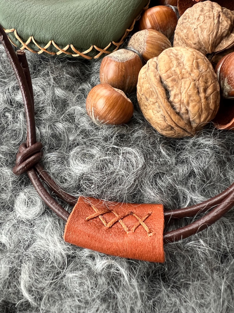 May include: A brown leather strap with a stitched brown leather flap. The strap is lying on a grey fluffy surface with a pile of hazelnuts and walnuts.