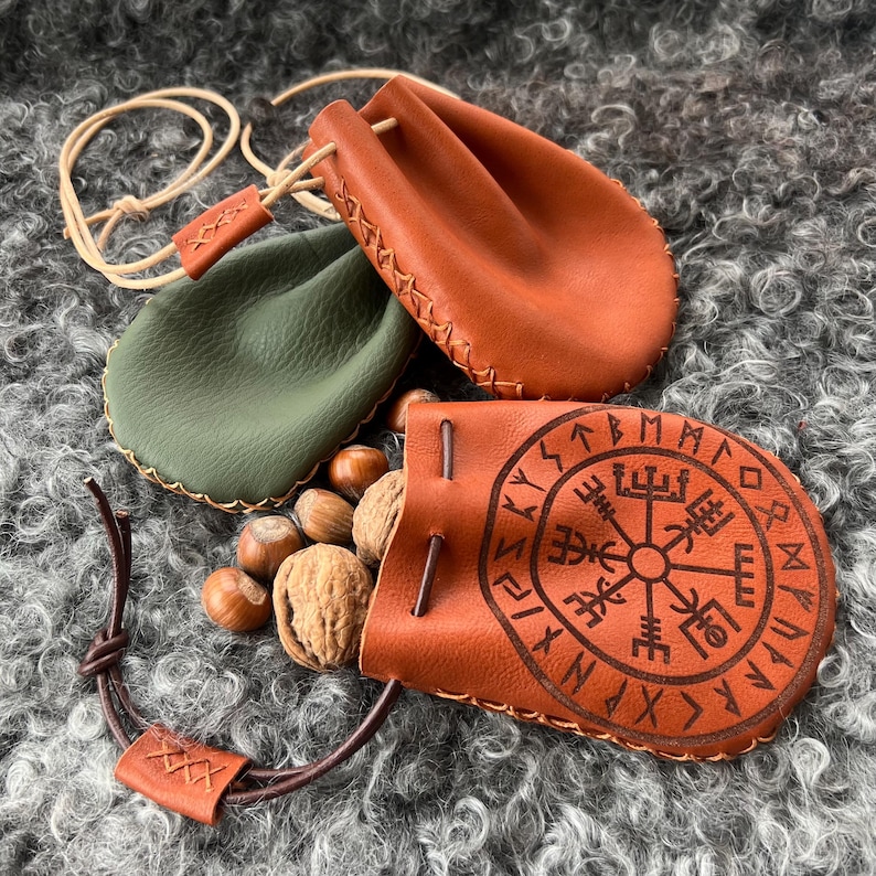 May include: Two small leather pouches with drawstrings. One pouch is green and the other is brown with a compass and runes design. Both pouches are on a fluffy brown surface.