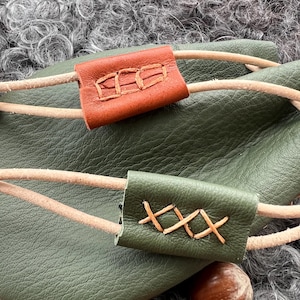 May include: A green leather bag with two brown leather straps. The straps have decorative stitching. The bag is sitting on a fluffy grey surface.