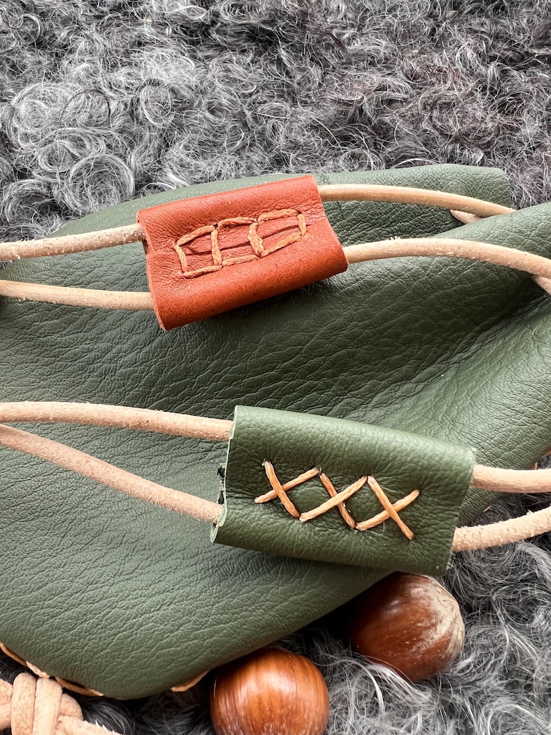 May include: A green leather bag with two brown leather straps. The straps have decorative stitching. The bag is sitting on a fluffy grey surface.