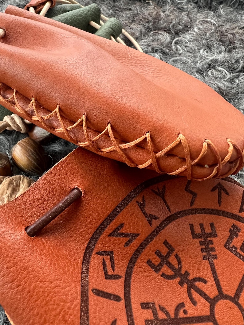 May include: A brown leather pouch with a braided leather strap and a decorative design of runes and symbols. The pouch is closed with a drawstring.
