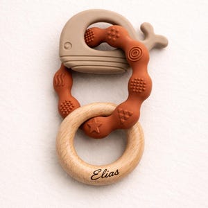 Könnte beinhalten: Ein Beißring für Babys bestehend aus einer beigen Walform aus Silikon, einem rostfarbenen, strukturierten Silikonring mit verschiedenen geometrischen Mustern und einem hellen Holzring mit der Gravur Elias in schwarzer Schreibschrift.