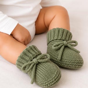 Könnte beinhalten: Ein Paar olivgrüne gestrickte Babyschuhe mit geripptem Bündchen und einer dekorativen Schleife auf der Vorderseite. Die Schuhe sind in einem strukturierten Strickmuster gefertigt und haben eine weiche, abgerundete Zehenpartie.