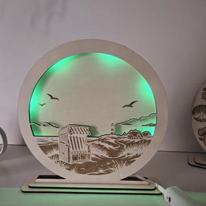 Può includere: Un espositore luminoso circolare in legno con una scena costiera tagliata al laser che mostra una sedia da spiaggia, un faro e uccelli. Il design ritagliato è illuminato da dietro con una luce verde brillante, creando un effetto silhouette.