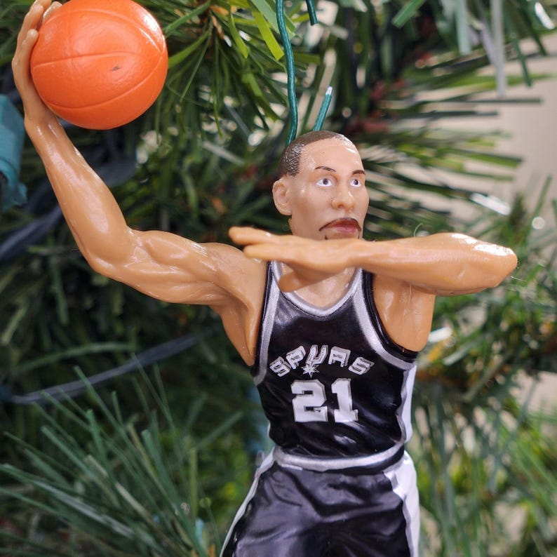 May include: A basketball-themed Christmas ornament depicting a basketball player in a black and white jersey with the number 21. The player is in mid-motion, holding an orange basketball. The ornament is hanging from a green string.