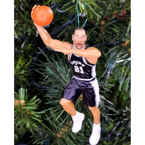 May include: A figurine of a basketball player in a black and white uniform, number 21, holding an orange basketball. The figurine is in a shooting pose, with white sneakers. The figurine is a Christmas tree ornament.