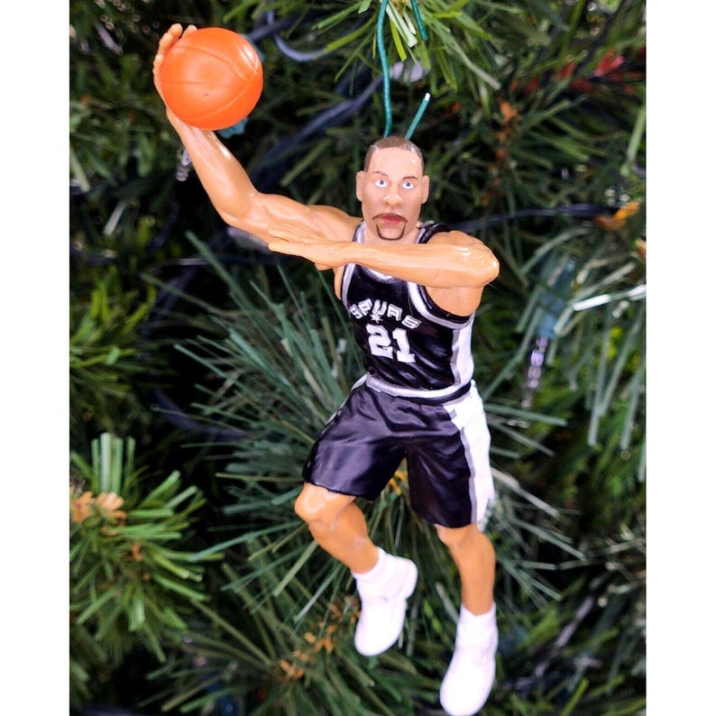 May include: A figurine of a basketball player in a black and white uniform, number 21, holding an orange basketball. The figurine is in a shooting pose, with white sneakers. The figurine is a Christmas tree ornament.