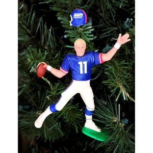 May include: A Christmas tree ornament depicting a football player in a blue jersey with the number 11, white pants, and a red football. A blue helmet with "Giants" is above the player. The ornament is on a green base.