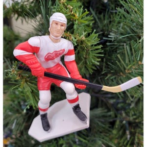 May include: A hockey player figurine in a red and white uniform, holding a hockey stick. The figurine is wearing a white helmet and black skates. The uniform features the Detroit Red Wings logo. The figurine is on a white base, and is a Christmas tree ornament.