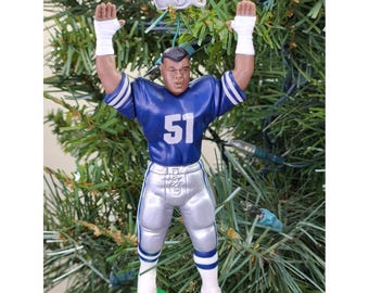 May include: A football player ornament in a blue jersey with the number 51, silver pants, and white socks. The ornament has a white helmet with a blue star and is posed with arms raised. The ornament is hanging on a Christmas tree.