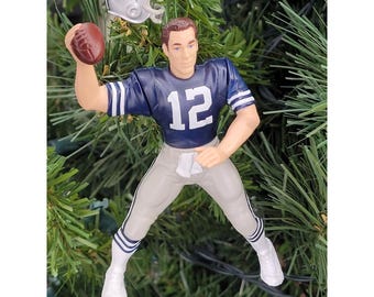 May include: A football-themed Christmas tree ornament. The ornament features a figurine of a football player in a blue jersey with the number 12, throwing a brown football. A silver helmet with a star is also included.