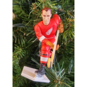 May include: A Christmas tree ornament depicting a hockey player in a red uniform with white accents and a white winged logo. The player is in a skating pose, holding a hockey stick. The ornament is attached to a white base.