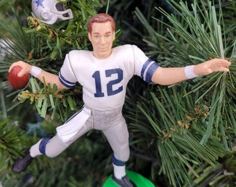 May include: A figurine of a football player in a white and blue uniform with the number 12, holding a red football. A miniature helmet with a star is above the figurine. The figurine is on a green base, set against a backdrop of green pine needles.