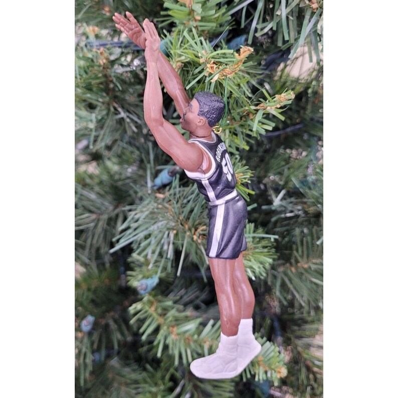 May include: A basketball player ornament in a black and white uniform, mid-shot, with arms extended upwards. The figure is set against a backdrop of green Christmas tree branches. The ornament is detailed with a jersey number and white sneakers.